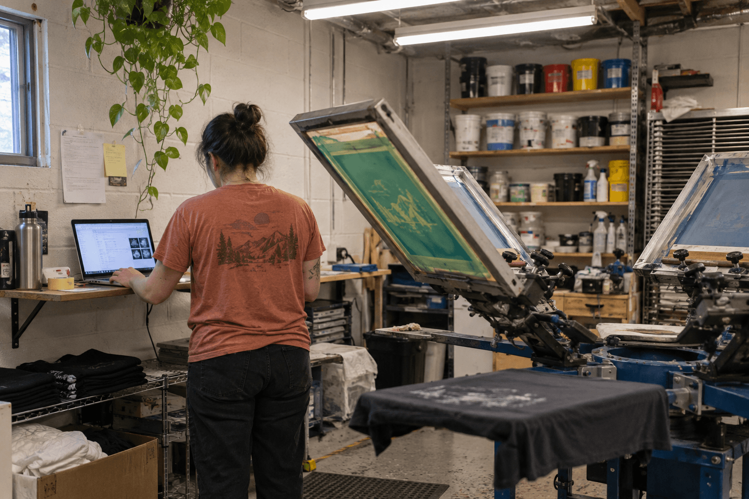 A small-business owner checking a laptop beside a printing press.